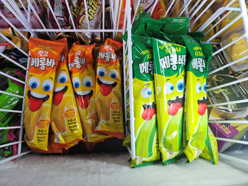편의점 냉동고 매대에 가득 찬 초록색 멜론맛과 주황색 망고맛 메롱바 아이스크림. 모두가 망설일 때 사장의 촉으로 한겨울에 물량을 선점하여 '오픈런' 열풍을 일으킨 신상 아이템의 실전 기록 사진.
