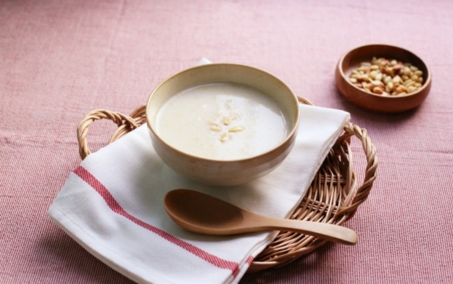 야생에서 채취를 한 잣을 갈아서 쌀과 함깨 만든 잣죽을 동그란 그릇에 담아서 붉은 색 테이블 위에 대나무로 만든 깔판 위에 놓아두고 그 옆에는 나무 숟가락과 잣을 담은 동그란 그릇을 놓아두고 찍은 사진