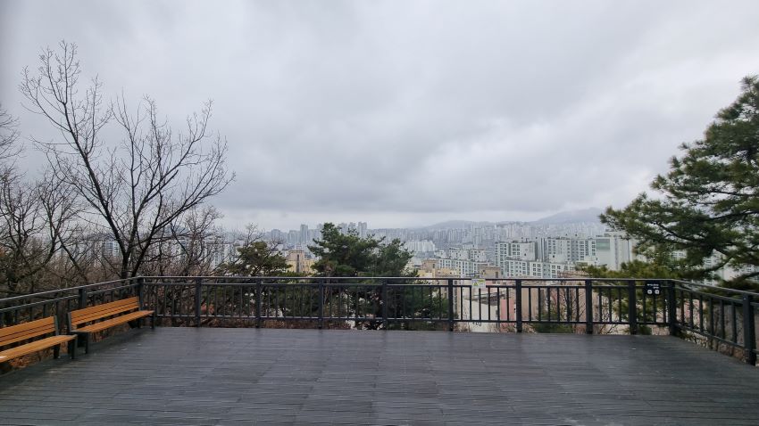 데크전낭대&#44; 둘러쳐진 안전난간&#44; 흐린 하늘&#44; 멀리 보이는 시가지 아파트 건물들&#44; 그 뒤로 북한산&#44; 우측에 큰 소나무&#44; 좌측에 벤치 둘&#44; 갈색칠&#44;
