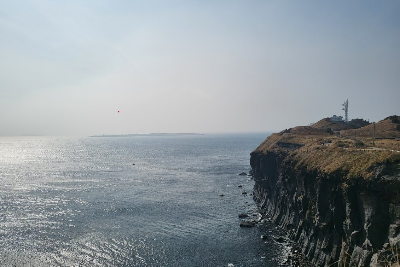 제주도의 바다. 오른쪽 절벽 밑으로 푸른 바다가 드넓게 펼쳐져 있다. 절벽에는 울타리와 건물들이 있다.
