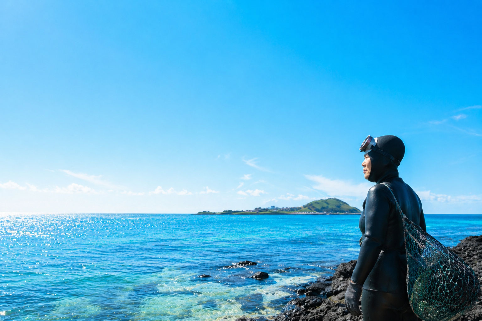 제주도의 푸른 바다를 배경으로 맑고 밝은 하늘 아래 해녀 한 명이 바다를 바라보며 서 있는 장면으로, 고요하고 평화로운 분위기를 담은 이미지