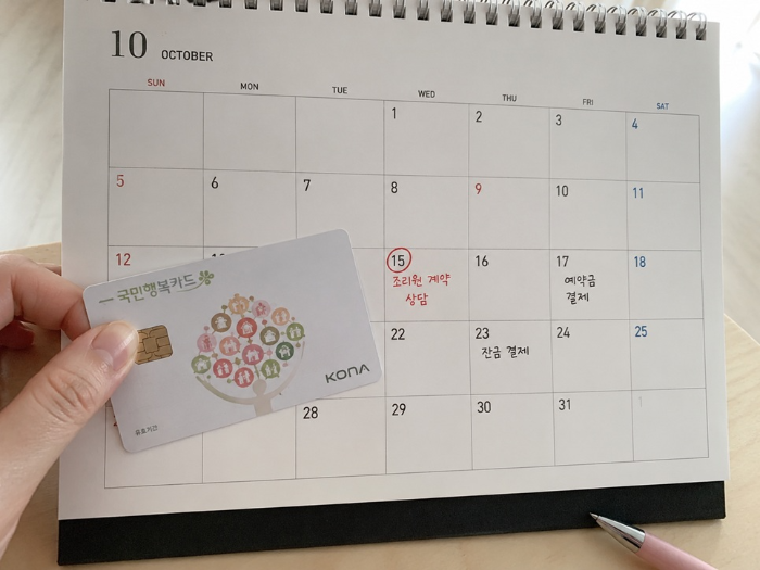 국민행복카드를 들고 탁상달력에 적힌 산후조리원 계약 상담, 예약금 결제, 잔금 결제 일정을 확인하며 첫만남이용권 바우처 사용 계획을 세우는 모습