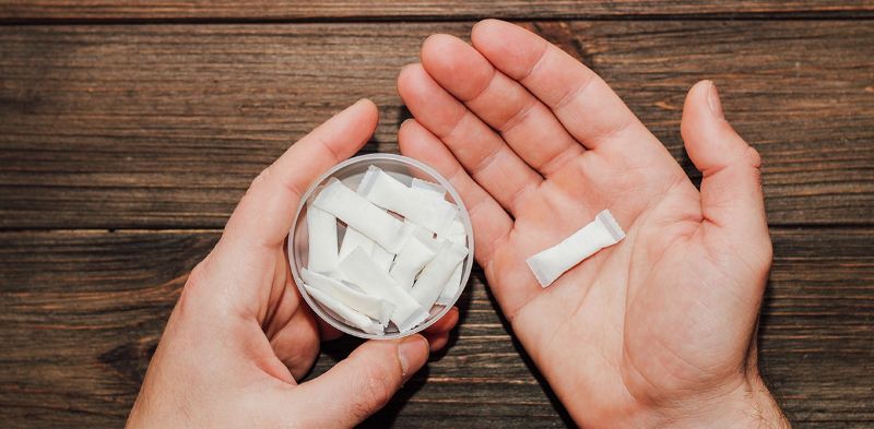 한 손에는 니코틴 파우치 통을, 다른 손에는 낱개의 파우치 제품을 들고 있는 모습. 흰색 주머니 형태의 니코틴 제품 구조를 보여주는 실제 사용 장면