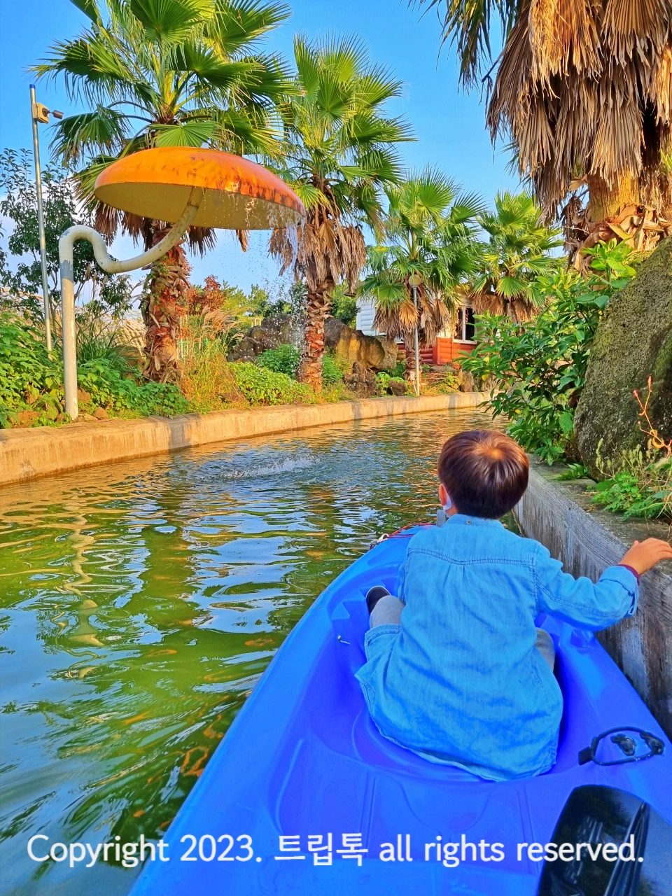 파란 카약을 탄 아이가 앞에서 버섯모양의 지붕에서 떨어지는 물을 보고 있다