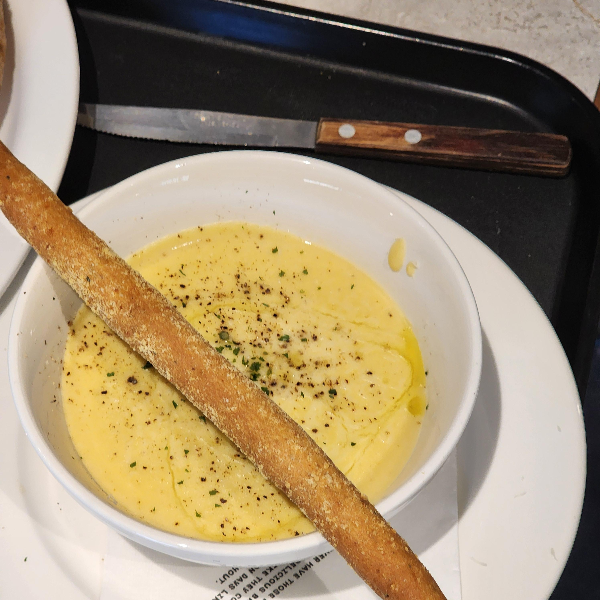 흰색 그릇에 담긴 크리미한 치즈 폴렌타. 위에는 후추와 허브가 뿌려져 있으며, 길고 바삭한 그리시니(이탈리아식 빵스틱)가 올려져 있음