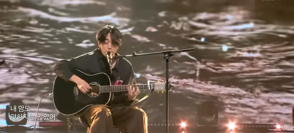 싱어게인3- 회색물결배경 검은 셔츠 가슴에 47번호표를 달고 검은색 기타를 치며 스탠딩마이크를 앞에 두고 땅을보고 의자에 앉아 노래하는 47호