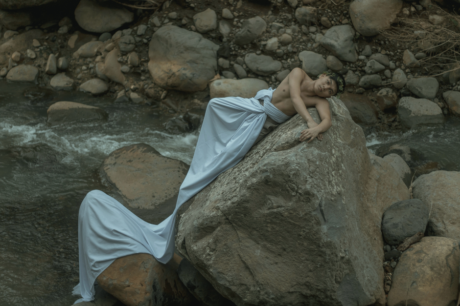 Male in white sheet and organic wreath looking at camera and lying on rough stone near foamy stream