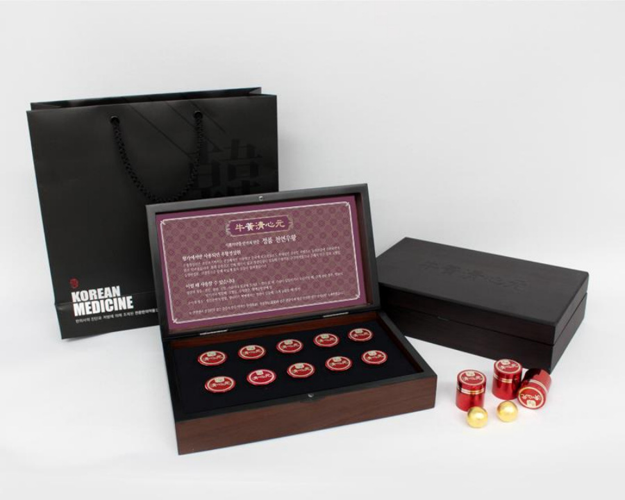 하얀 바닥 위에 한약을 담는 종이 가방과 그 앞에는 우황청심환을 담은 나무상자를 2개 두고, 하나는 뚜껑을 열어서 속에 보이게, 하나는 뚜껑을 닫은 상태로 두고, 그 앞에 우황청심환 2개와 우황청심환을 담은 빨간 통을 3개 놓아두고 찍은 사진