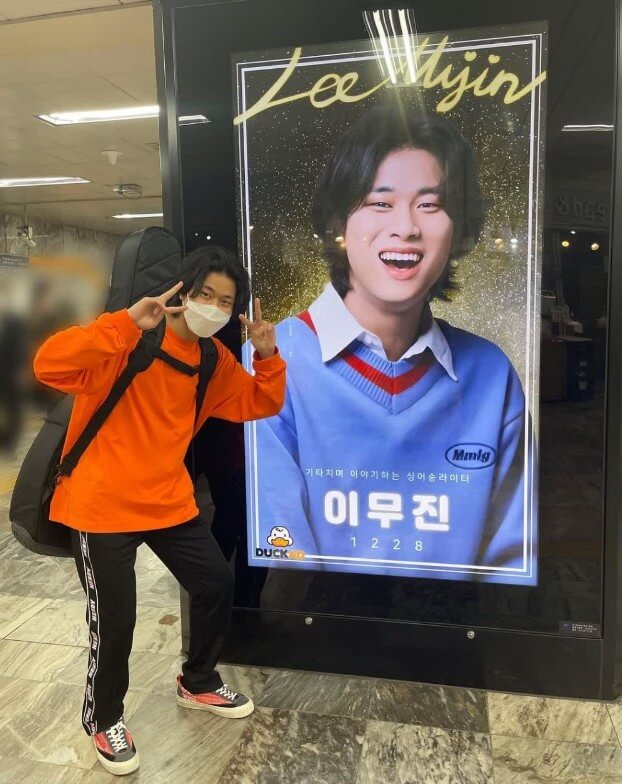 가수-이무진이-통기타를-등에-메고-자신의-사진-앞에서-양손으로-V를-그리며-서-있는-사진