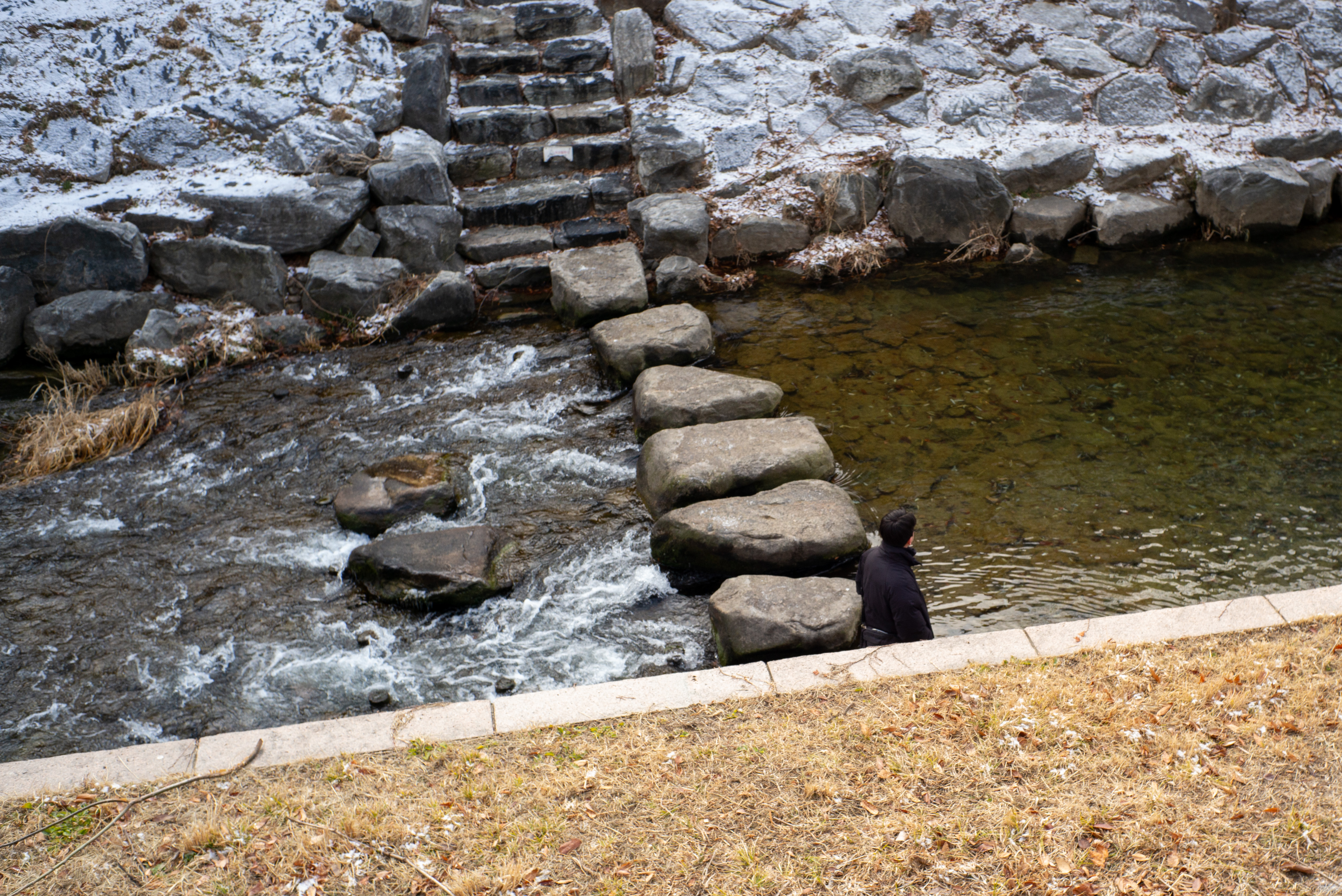 물이 흐르는 냇가에 놓인 돌 징검다리와 그 옆에 서서 물을 바라보는 사람의 모습