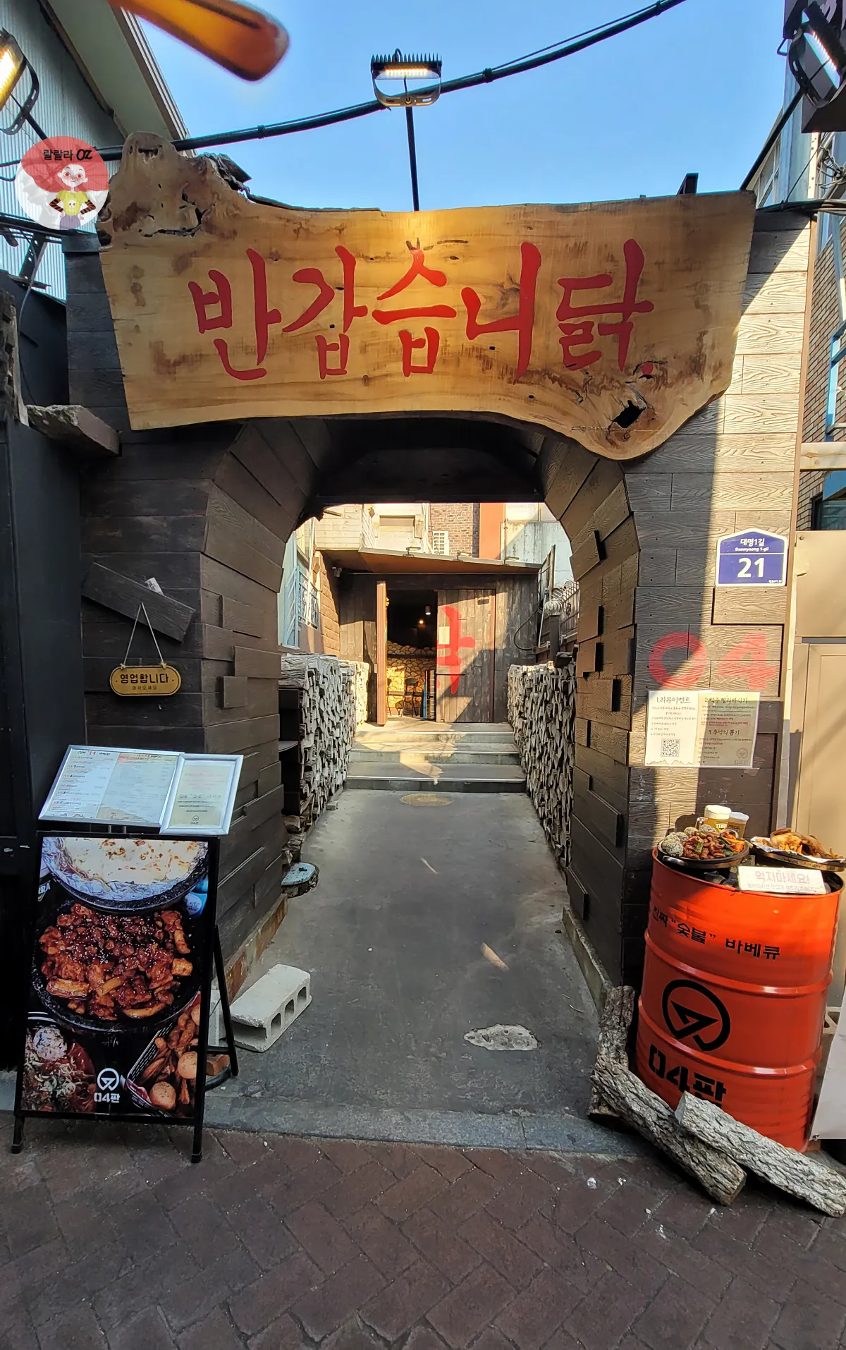 대학로 04판, 시골풍 닭갈비 식당 입구. 나무 아치형 간판에 &lsquo;반갑습니닭&rsquo;이라는 붉은 글씨가 걸려 있고, 양쪽에는 장작더미가 쌓여 있으며 메뉴판과 음식 모형이 전시된 아늑한 분위기