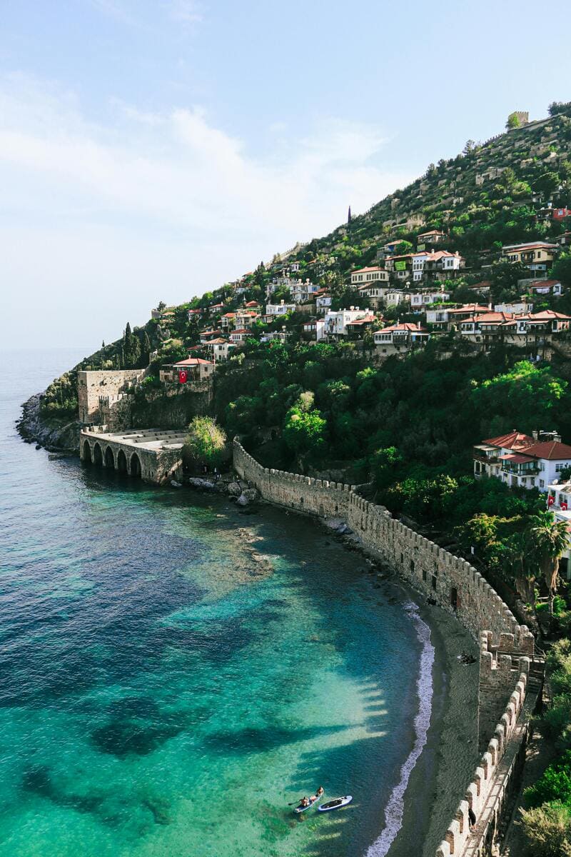 안탈리아 알라냐 지역의 성벽이 바다를 따라 뻗어 있으며, 언덕에는 붉은 지붕의 주택들이 계단식으로 빽빽하게 자리잡고 있습니다. 에메랄드빛 바닷가에서는 두 사람이 패들보드를 즐기고 있습니다