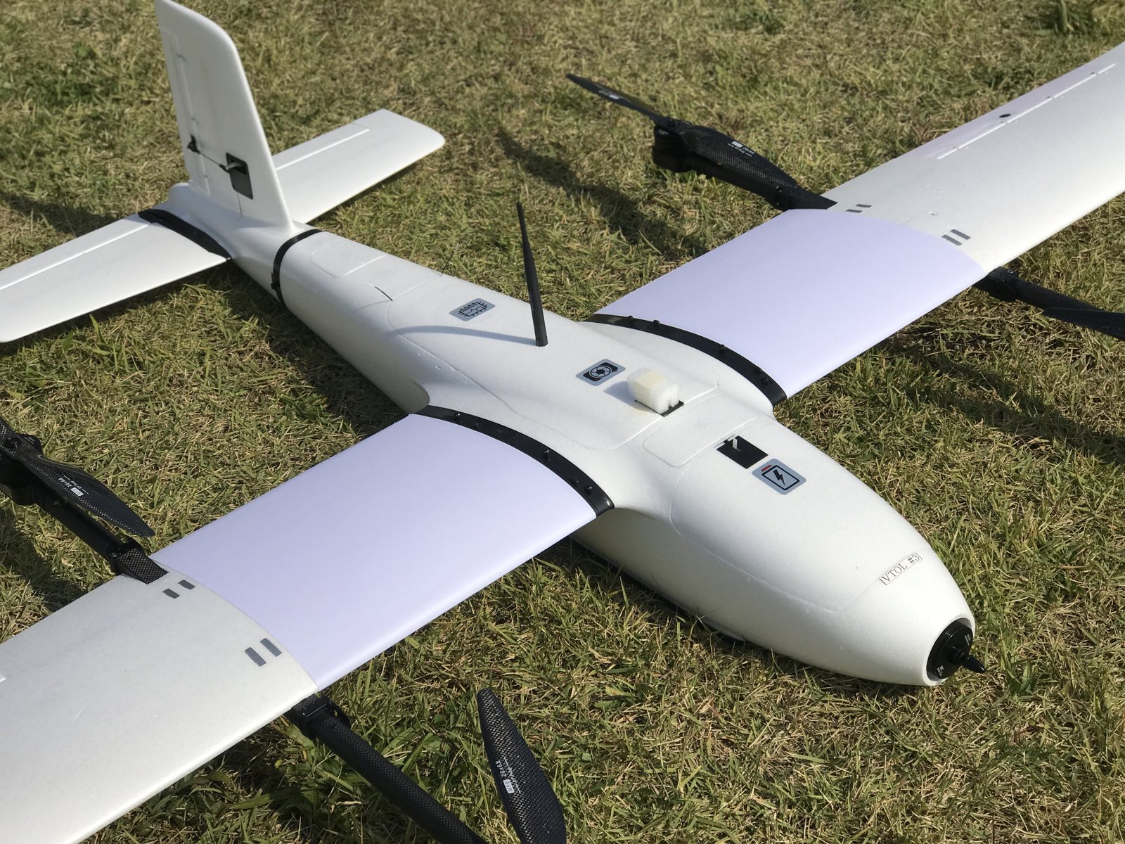 잔디 위에 놓인 흰색 VTOL 기체, 날개 윗면 일부에 연보라색 필름이 보인다