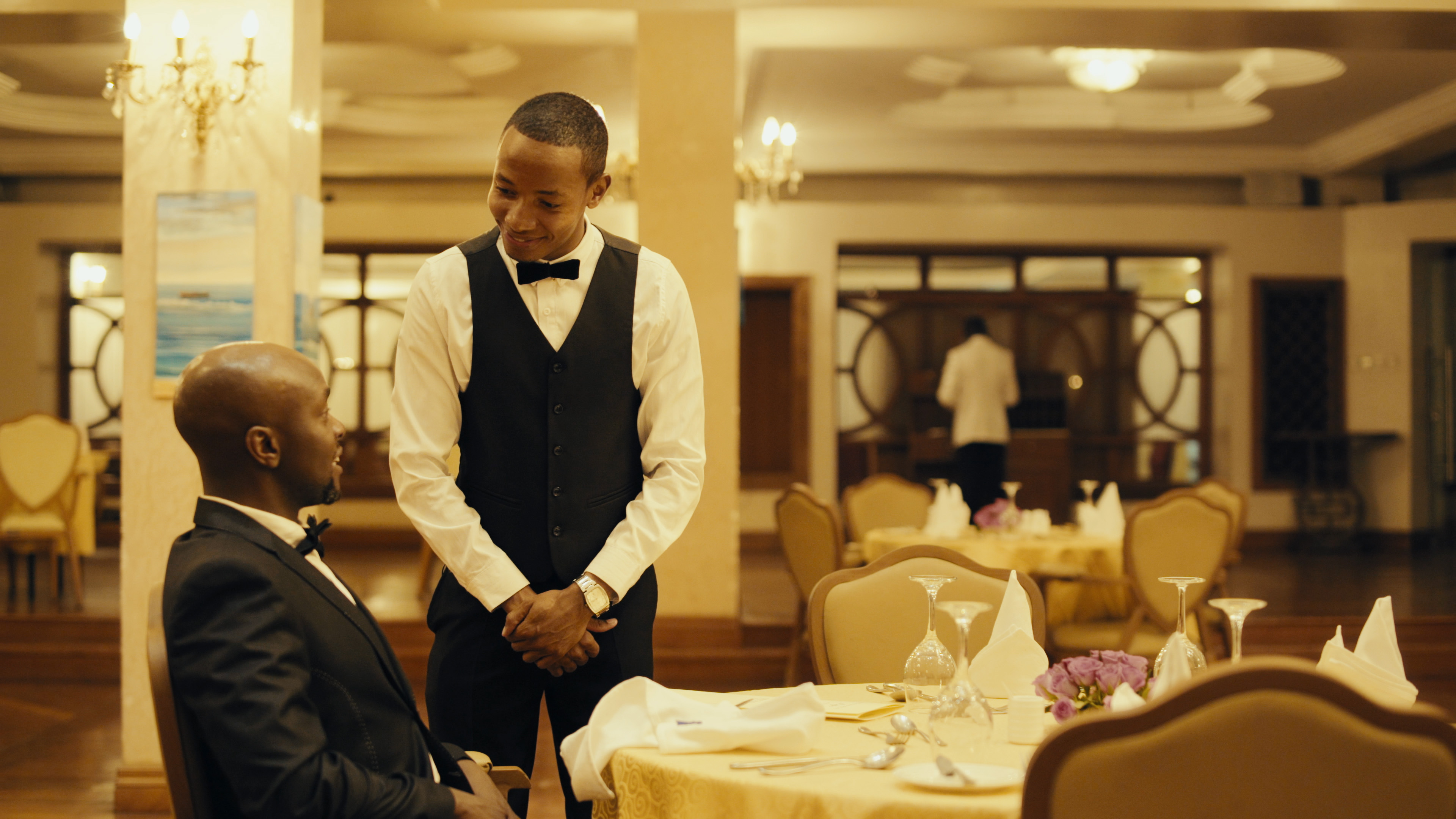 영화 나이로비에서의 외식 가이드 스틸컷, 웨이터와 손님이 고급 레스토랑에서 마주한 장면으로 10달러의 꿈과 도시의 불평등을 상징함