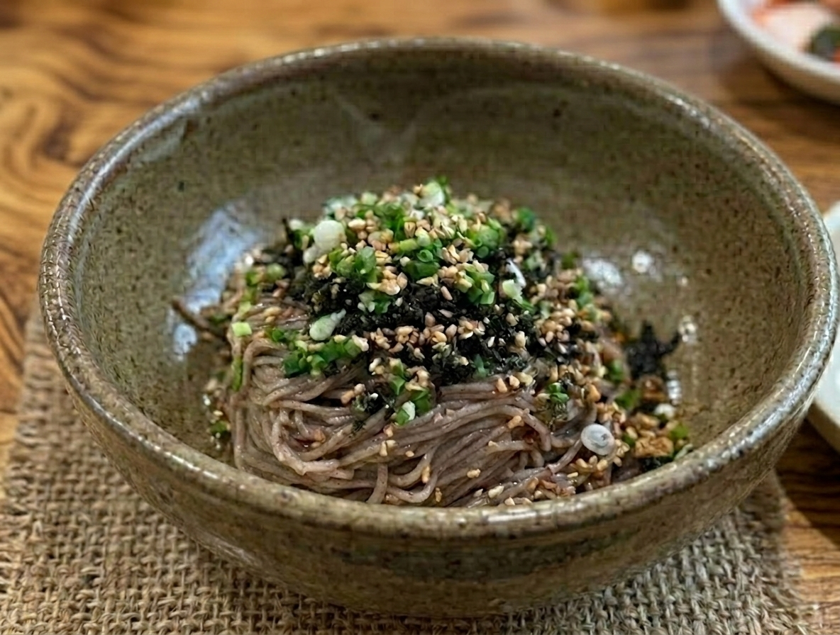 나뭇결이 살아있는 원목 테이블 위, 삼베 식탁 매트와 도자기 그릇에 정갈하게 담긴 들기름 막국수.