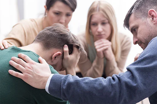 머리를-감싸고-고개를-숙이고-있는-남성을-세사람이-위로하고-있음