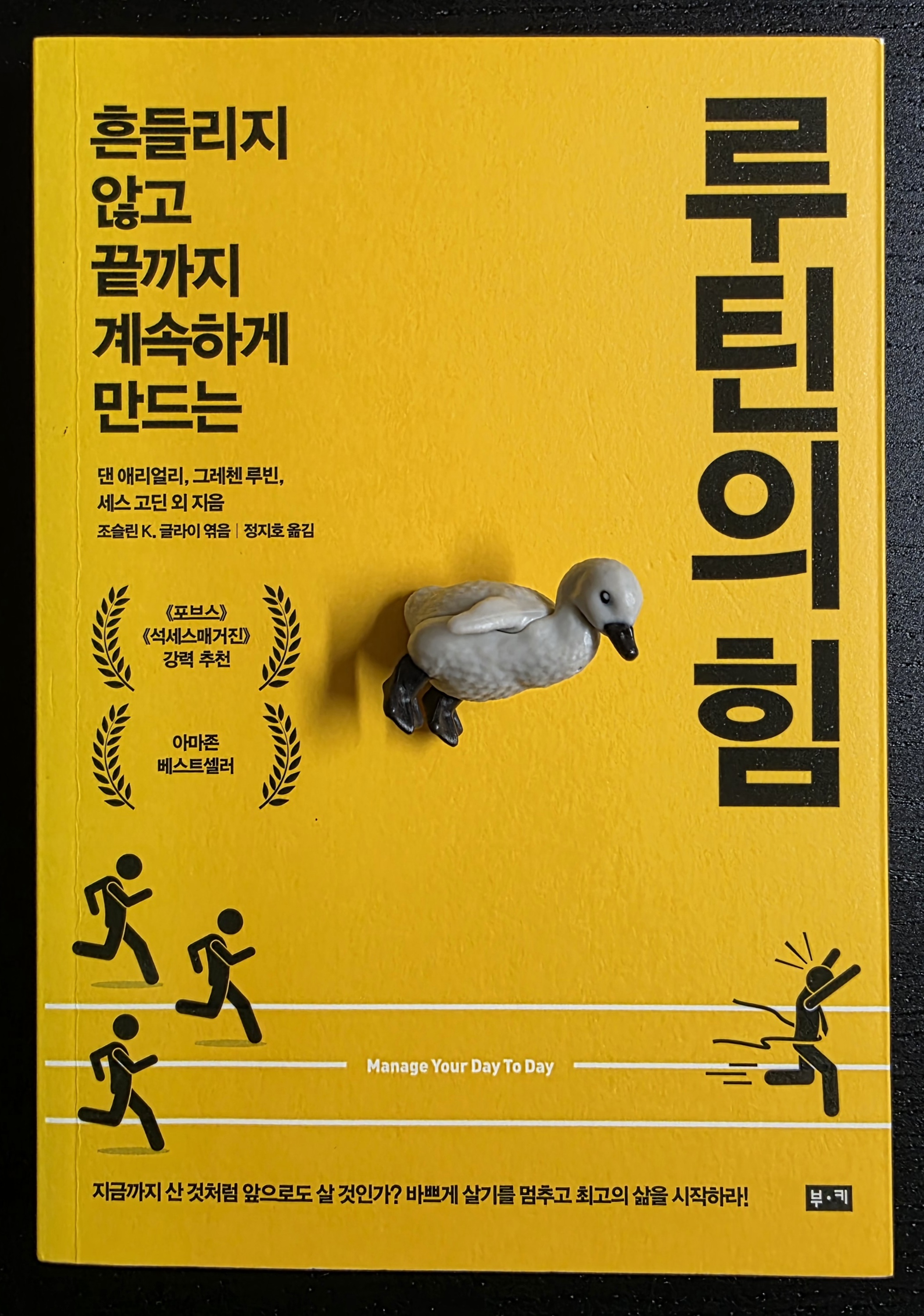 노란색 표지 위를 걷는 듯한 펭귄 피규어와 루틴의 힘 책