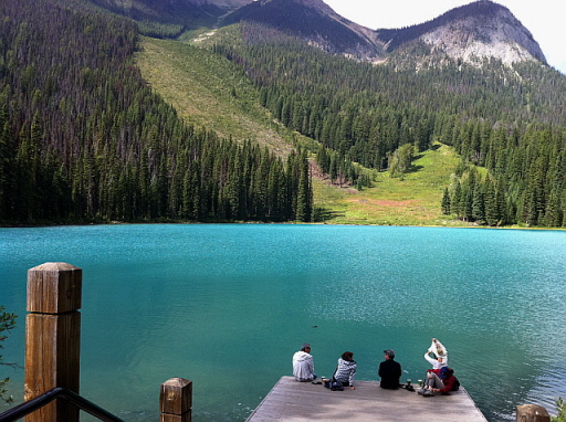 에메랄드빛 호숫가 나무 데크에 앉아 있는 사람들 뒤로 펼쳐진 캐나다 로키 산맥 풍경