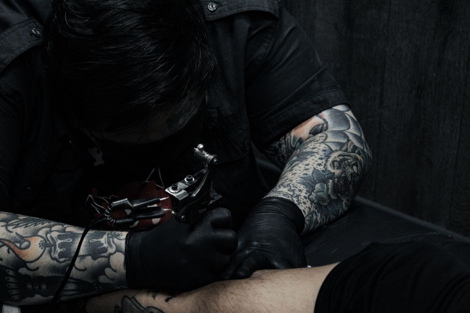 Tattoo artist focused on creating an intricate arm tattoo with a tattoo machine in a dimly lit studio.