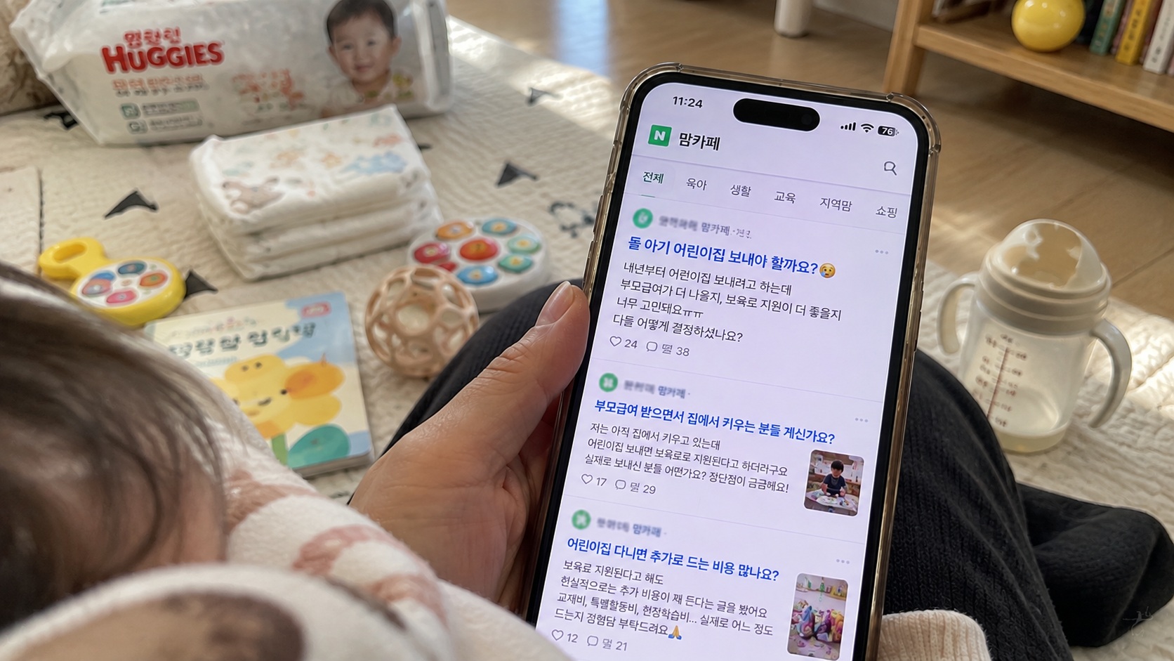 아기 장난감과 기저귀가 놓인 거실에서 스마트폰 맘카페를 보며 부모급여와 어린이집 보육료 지원을 고민하는 모습