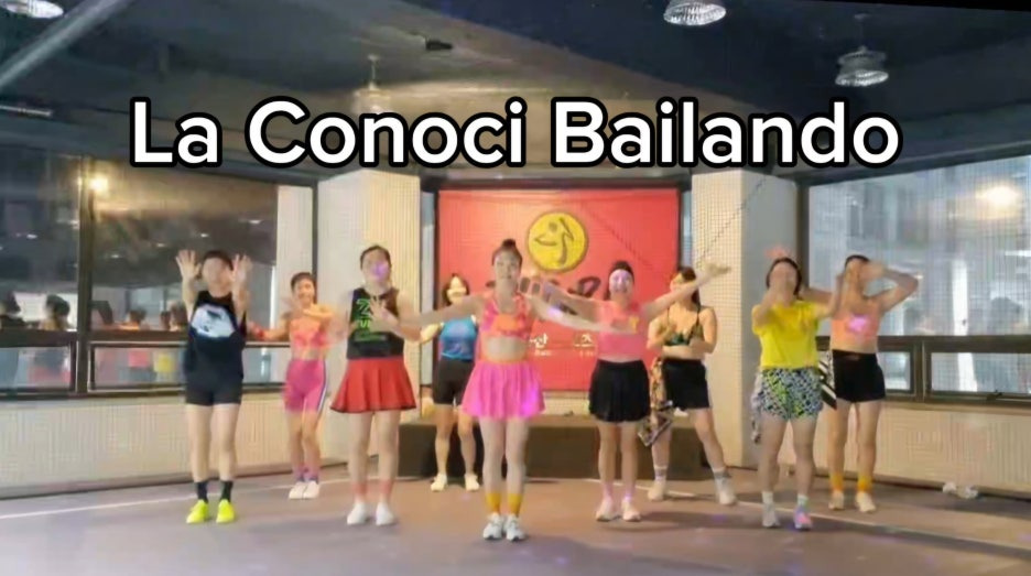 ZIN 해나 강사의 둔산줌바 수업 중, 'La Conoci Bailando' 음악에 맞춰 열정적으로 단체 안무를 선보이는 회원들. 형형색색의 밝은 의상을 입고 환한 표정으로 신나게 춤을 추는 모습이며, 배경에는 큰 붉은색 ZUMBA 배너가 걸려 있다