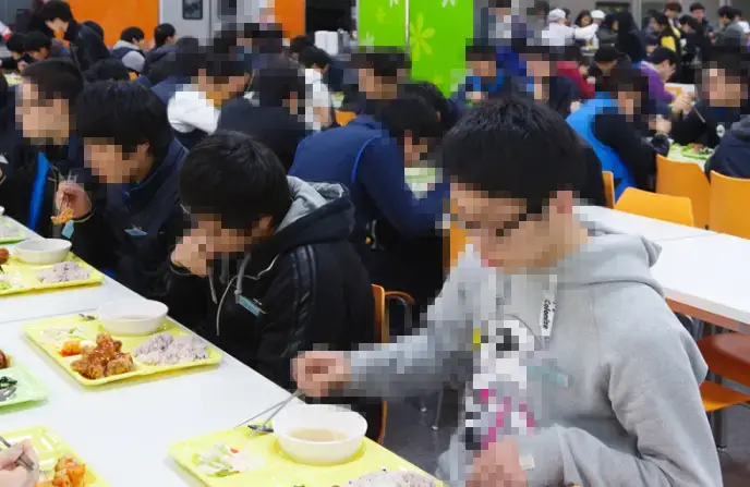 재수학원- 단체식당 안 흰색 긴테이블에 줄지어 앉아 노란 식판에 담근 음식을 먹고 있는 학생들