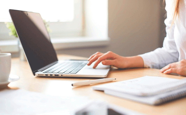 책상에 노트북이 있고 하얀 블라우스를 입은 여성이 손으로 노트북 키보드를 치고있다.