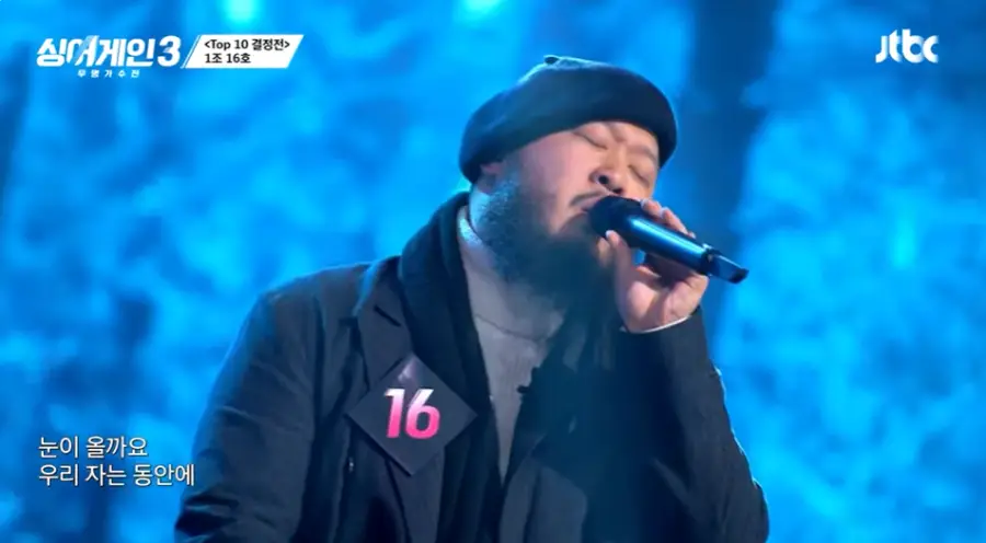 싱어게인3-
파란배경 검은색 외투에 회색 폴라티 검은 배레모에 검은 마이크를 들고 노래하는16호의 오른쪽 모습