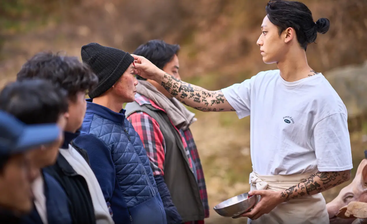 파묘- 흰옷을 입고 왼손에 스텐그릇 오른손으로 남자들의 이마에 손을 대고 있는 봉길과 맞은편의 5명의 사람들