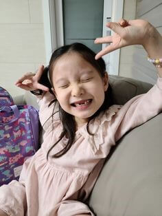 분홍옷을-입은-여자아이가-윗니-두개가-빠진채로-소파에앉아-V를-그리며-환하게-웃는-모습