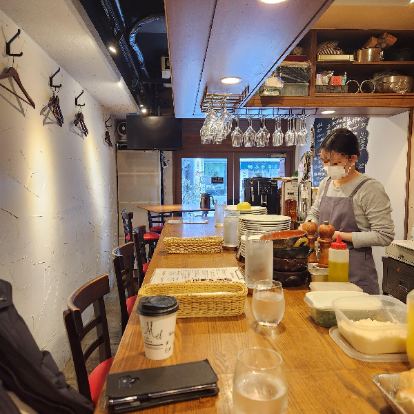 흰 벽에 좁은 공간. 바형 테이블이 일자로 배치되어 있다. 음식을 만드는 직원이 마스크를 쓰고 있다.