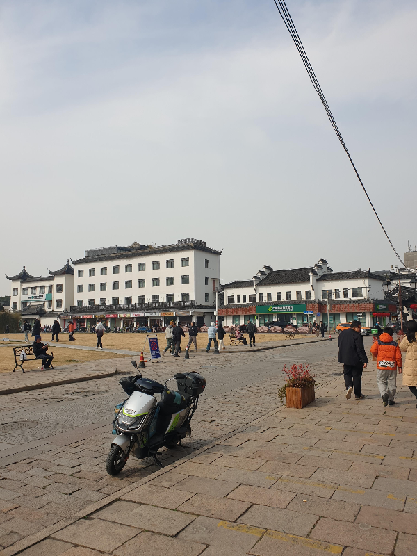 넓은 공터에 사람들이 있고 그 뒤로 흰색 2층과 4층 건물이 보인다.