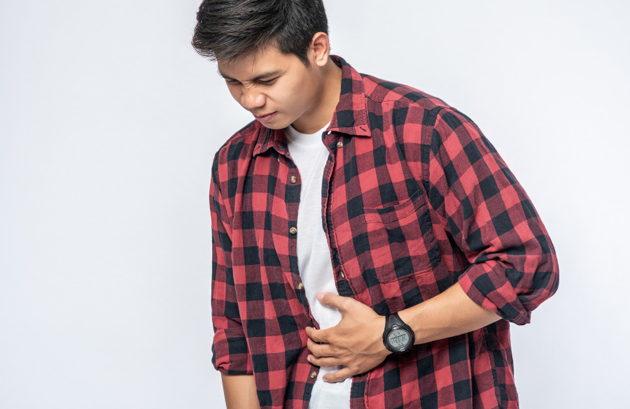 붉은색의 체크 무늬 셔츠를 입은 젊은 동남아 남성이 위장의 통증 때문에 왼쪽 손으로 배를 만지며 콧등을 찡그리며 통증을 견디고 있는 것을 찍은 사진