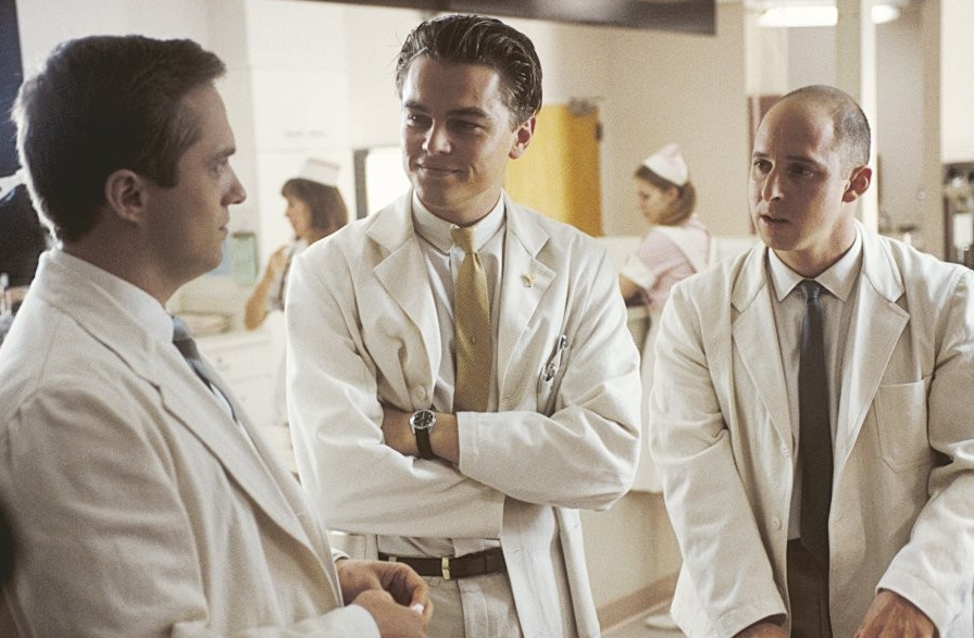 병원 복도에서 흰 가운을 입은 남자가 팔짱을 낀 채 동료들과 대화를 나누고 있는 장면