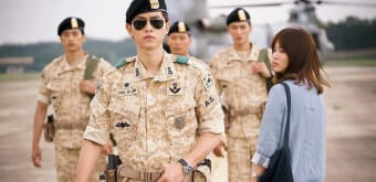 태양의 후예 남주인공이 군복을 입고 동료들과 함께 걸어오고 있고, 여주인공이 그 모습을 뒤돌아 보고 있다.