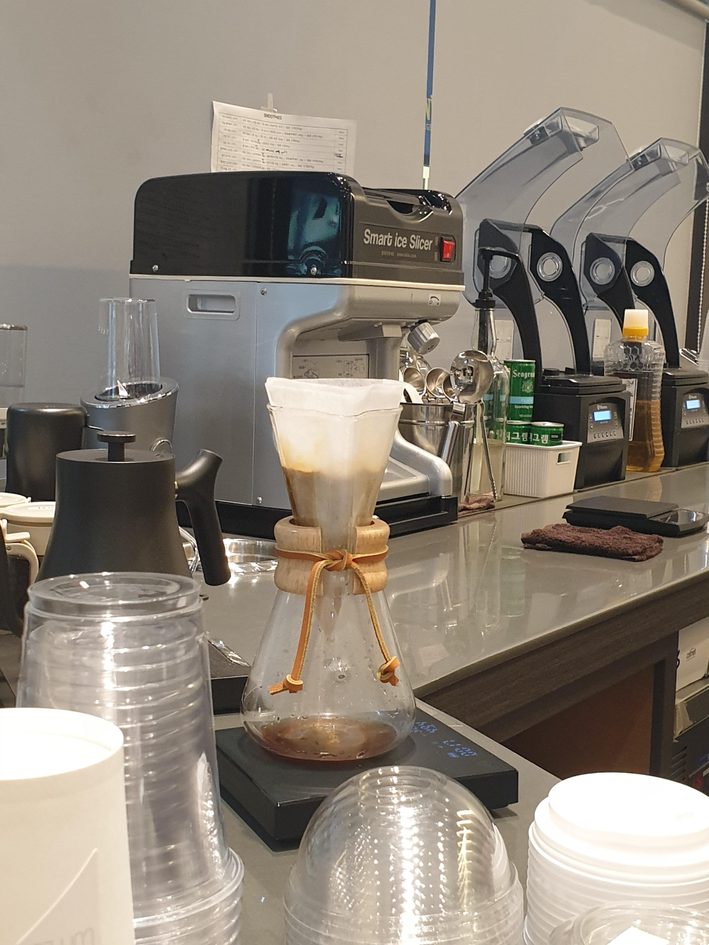 케멕스 유리 드리퍼에 커피 파우더가 담겨 있다. 뒤쪽에는 블렌더들이 보인다.