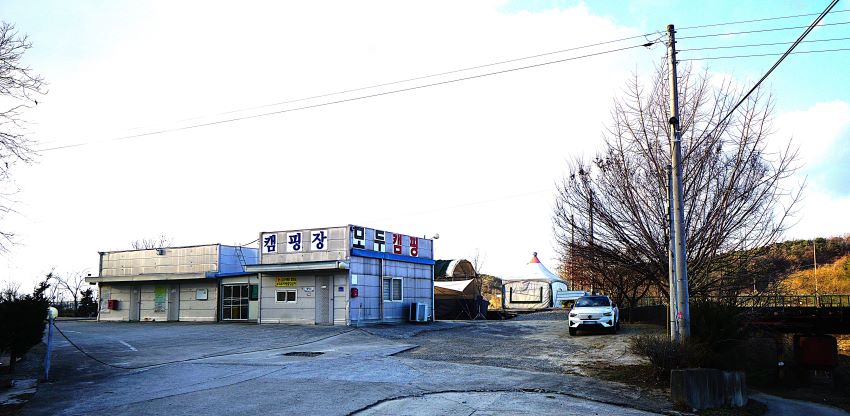 푸른하늘, 단층 건물 두 동 윗면 벽에 '캠핑장, 모두캠핑'이라고 쓰인 큰 글씨, 우측에 주차된 흰 승용차, 승용차 뒤에 흰 몽고텐트, 우측에 개울,