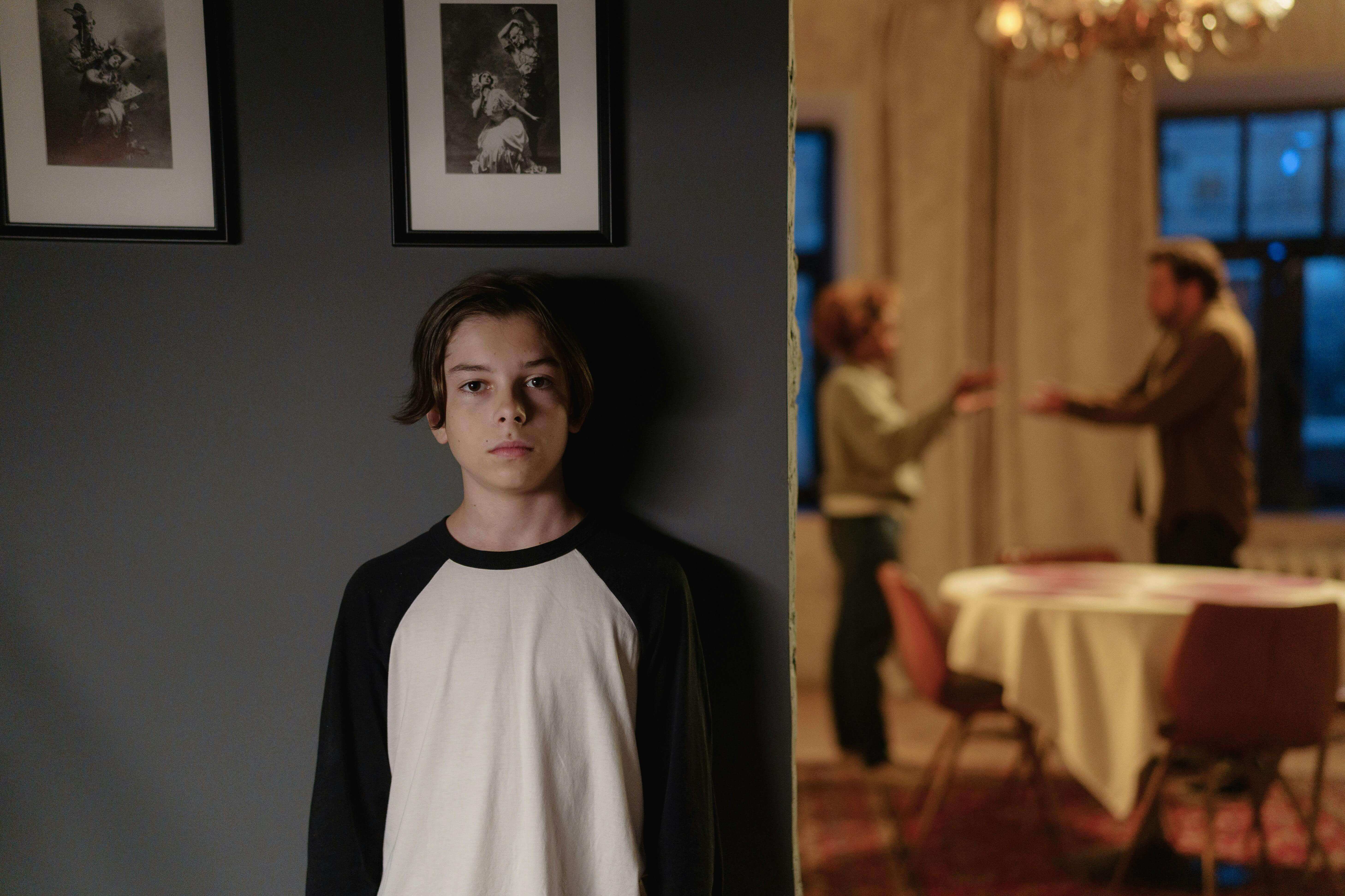 거실 한켠에서 부모의 다툼을 지켜보며 혼자 서 있는 무표정한 청소년