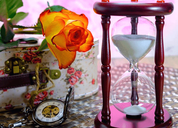 하얀색 모래가 담긴 모래시계가 있다. 왼쪽에는 주황색 장미와 둥그런 시계가 있다.