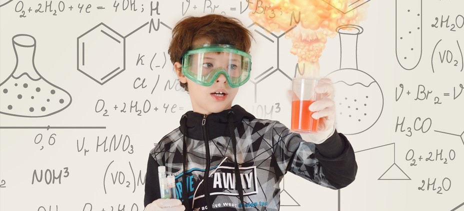 과학자 소년이 어려운 과학 공식이 가득 쓰여 있는 칠판 앞에서 실험을 하고 있는 사진