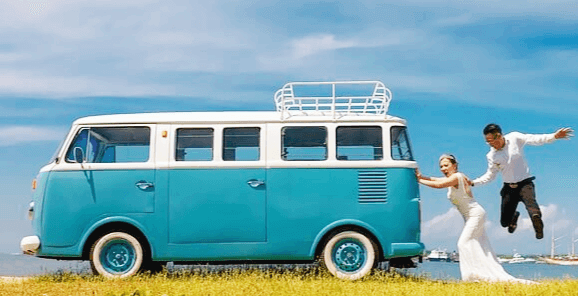 파란 승합차를 뒤에서 밀고있는 웨딩드레스를 입은 신부와 폴짝 뛰고있는 신랑이 있다.