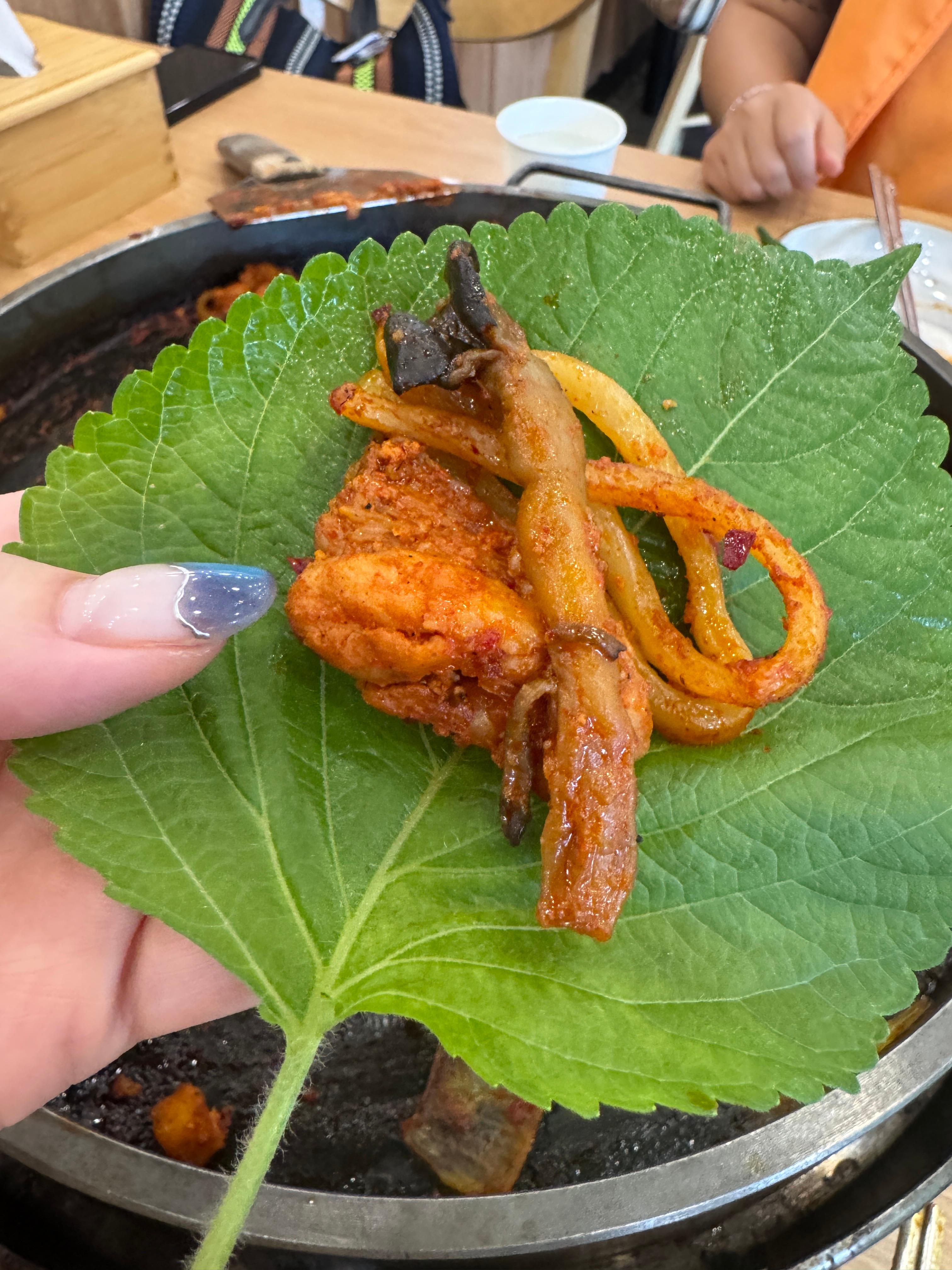 깻잎 위에 닭갈비&amp;#44; 우동사리&amp;#44; 버섯을 함께 올린 쌈. 뒷배경에는 닭갈비가 거의 비워진 철판이 보인다. 