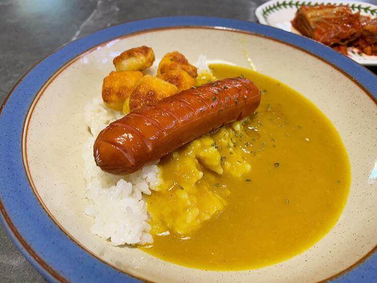 파란색 테두리가 있는 접기에 왼쪽에는 밥, 오른쪽에는 카레가 담겨져 있다. 밥 위에서 칼집이 난 소세지 한 개와 팝콘 치킨 4 개가 있다. 접시 뒤로 하얀 접시에 담긴 김치가 있다