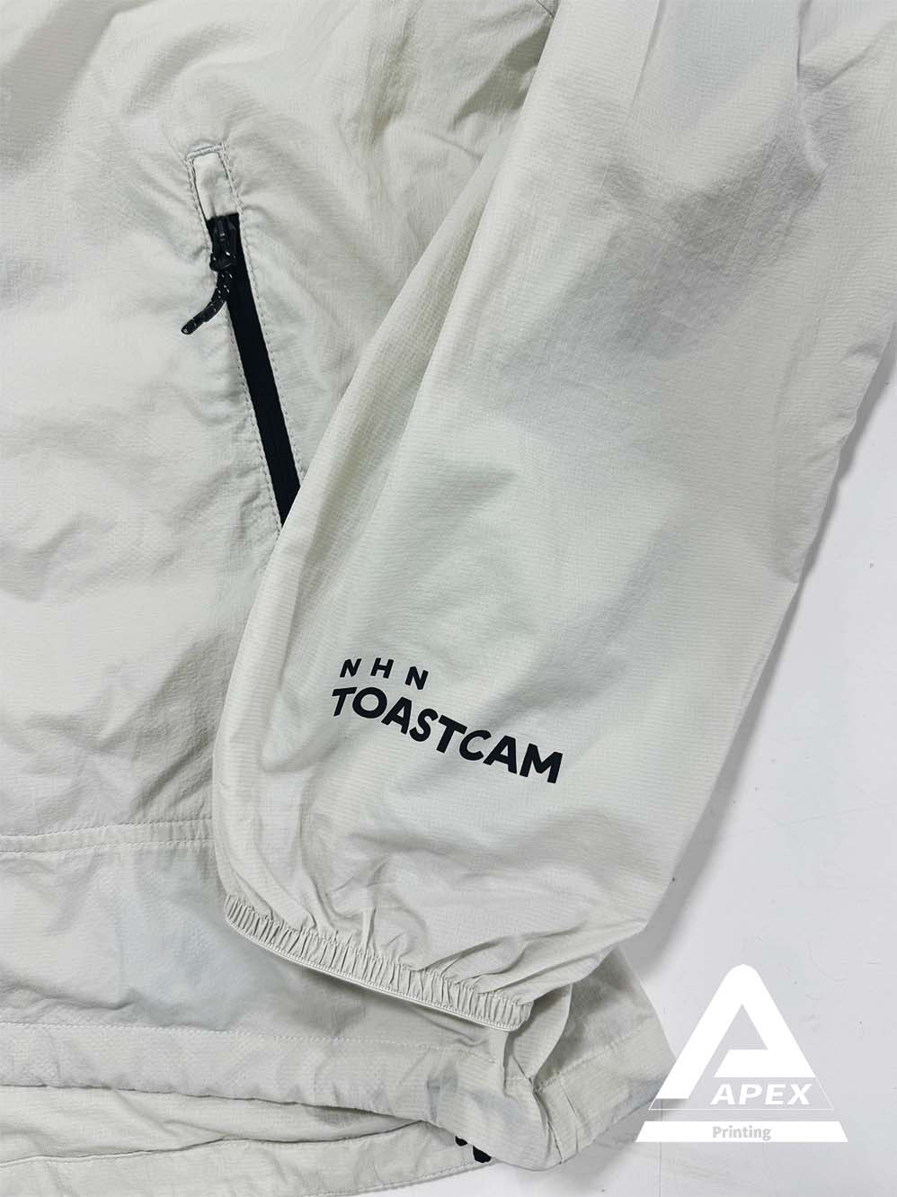 뉴발란스 연베이지 바람막이의 소매 부분 확대 이미지로, &quot;NHN TOASTCAM&quot; 로고가 깔끔하게 프린팅된 모습이 보임.