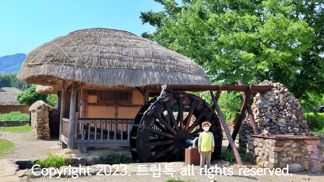 초가집 옆에 물레방아가 있고 그 앞에 남자아이가 서있다.