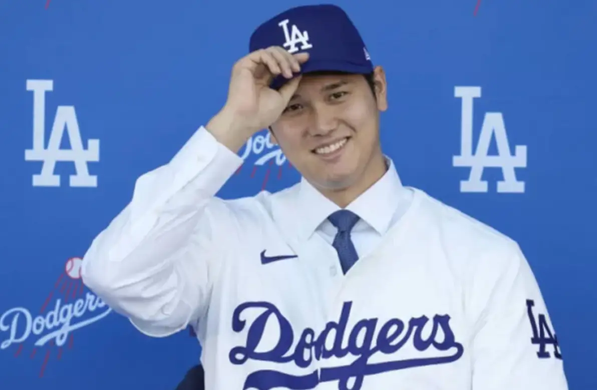 파란색 원단 위 흰글씨 LA dodgers 앞 흰유니폼 파란 야구모자 챙을 오른손으로 잡고 미소짓는 오타니