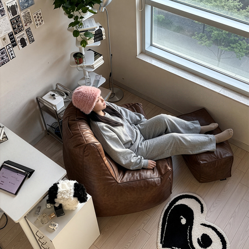 창가 책상 옆 방에서 분홍 모자를 쓴 여성이 플래지어 톰빈백 하이백 브라운 기능성 가죽 빈백에 기대고 스툴에 발을 올린 모습을 위에서 본 사진