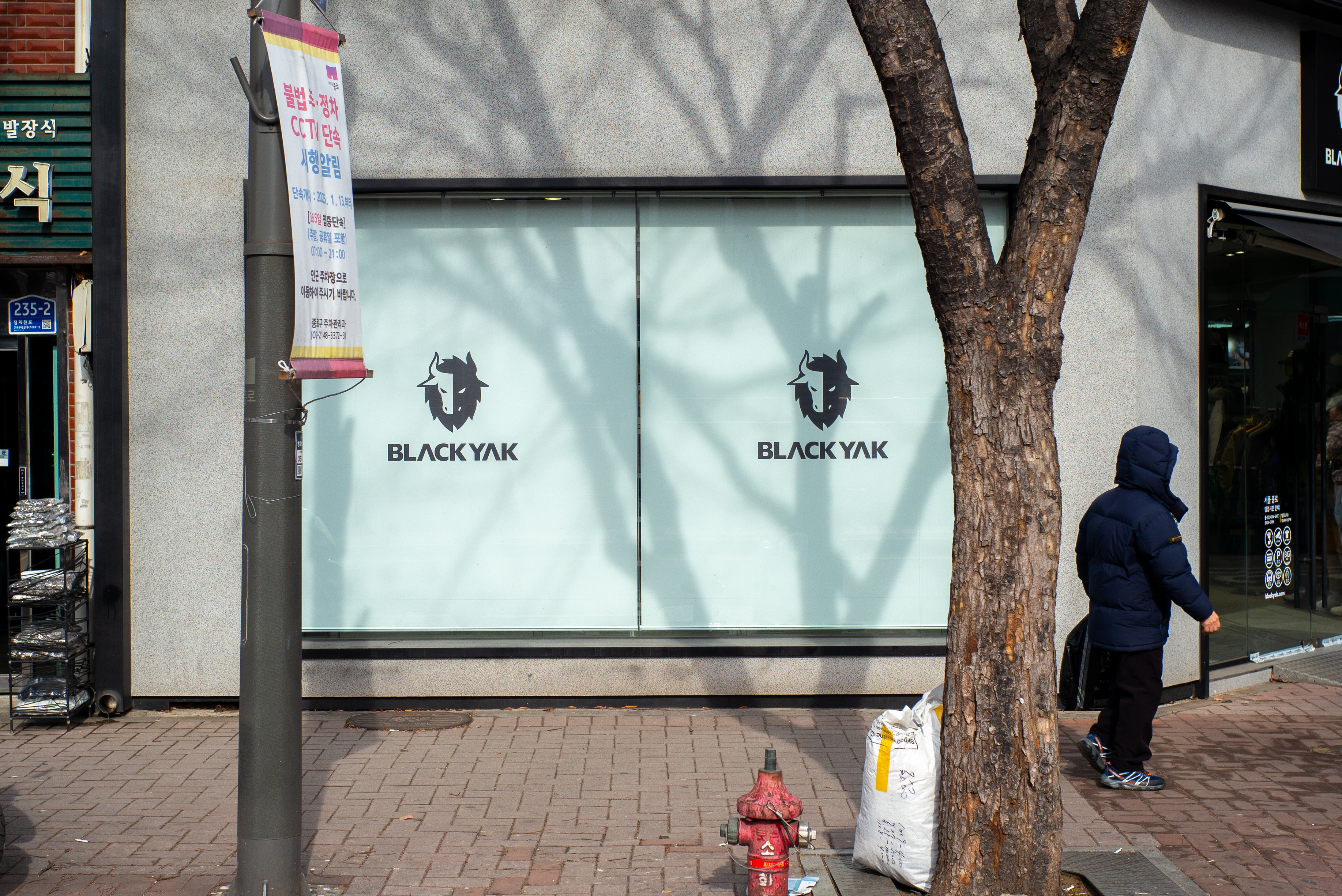 블랙야크 로고가 그려진 매장 유리창과 그 앞으로 드리워진 나무 그림자, 길을 지나가는 행인