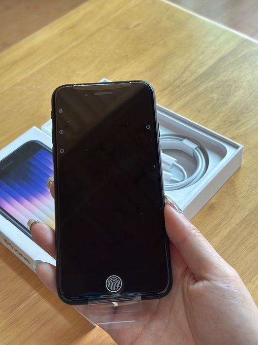 아이폰 SE3 전면 디스플레이를 손에 들고 있는 모습&amp;#44; 아래에는 지문인식 홈버튼 아이콘이 보이며 박스 안 구성품이 함께 놓여 있는 장면