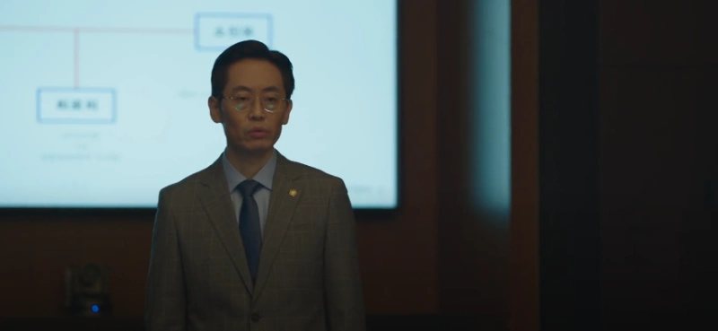 어느 고급스러운 회의실에서 단정히 넘긴 머리와 안경을 썼으며, 회색 정장을 입고 브리핑 중인 드라마 유괴의 날 박정훈 캐릭터